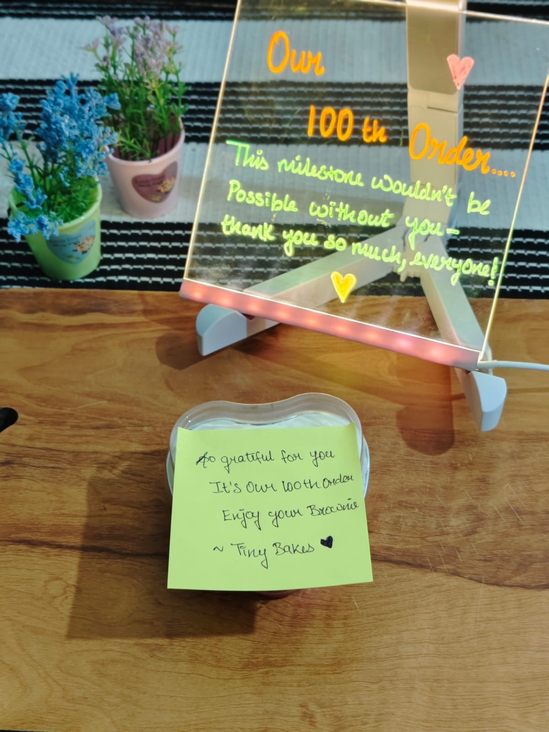 A glowing hand-lettered board celebrating Tiny Bakes' 100th order, beside a hand-written thank-you note